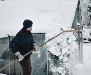 Man using a roof rake to remove snow from a greenhouse roof during a cold winter day in Estonia.
